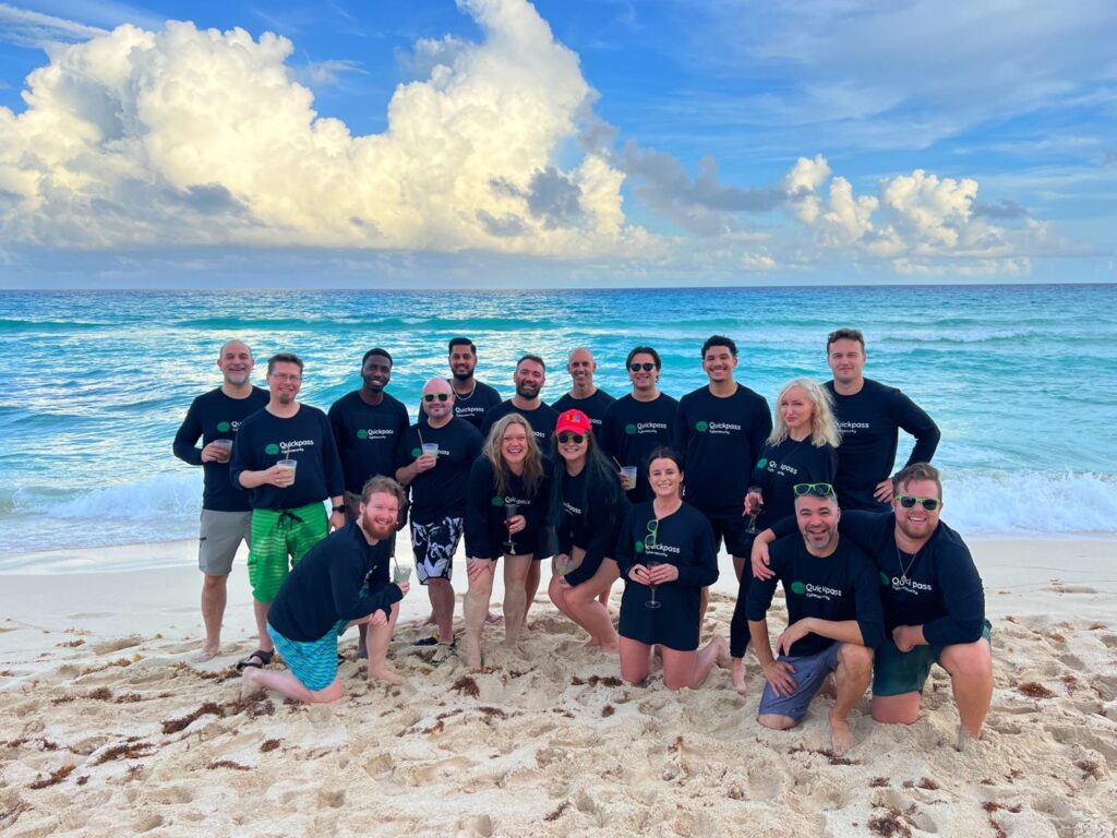 Group of people wearing matching shirts, smiling and posing on sandy beach in front of ocean.